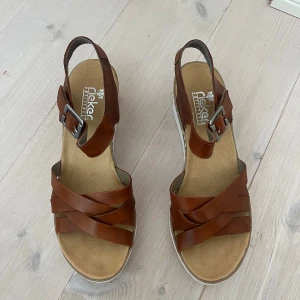 Bruna sandaler med kilklack från Rieker - Snygga sandaler från Rieker i brunt skinn med korslagda remmar och justerbart spänne vid ankeln. Skorna har en vit plattformsula och kilklack med korkdetalj, vilket ger en modern och bekväm look. Perfekta för varma dagar och sommarens outfits.