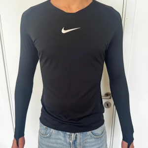 Svart långärmad Nike träningströja - Svart långärmad träningströja från Nike med vit logga på bröstet. Slimmad passform och tillverkad i ett stretchigt syntetmaterial som andas. Perfekt för dig som gillar sportig och clean stil.