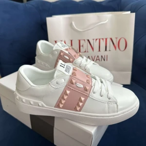 Valentino Rockstud sneakers vit/rosa - Snygga Valentino Garavani Rockstud sneakers i vitt skinn med ljusrosa band och ikoniska nitar. Klassisk rund tå, platt sula och vita snören. Perfekta för dig som vill ha en lyxig och trendig look med extra edge.