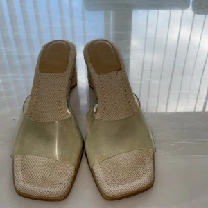 Beiga sandaler med kilklack - Snygga sandaler med transparent rem över foten och beige textil på sulan. Skorna har en hög kilklack i flätad jute och fyrkantig tå. Perfekta för sommarens alla tillfällen när du vill ha en stilren och trendig look.