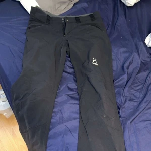 Svarta täckbyxor från Haglöfs - Svarta täckbyxor från Haglöfs med dragkedja och knappstängning framtill. Byxorna har bälteshällor och är perfekta för vinterns utomhusaktiviteter. Snygg och funktionell design med Haglöfs-logga på låret.