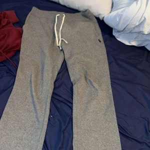 Grå mjukisbyxor från Polo Ralph Lauren - Grå mjukisbyxor från Polo Ralph Lauren med vit snörning i midjan och klassisk logga på höften. Byxorna har rak passform och är tillverkade i mjukt bomullsmaterial, perfekta för chill dagar eller träning.