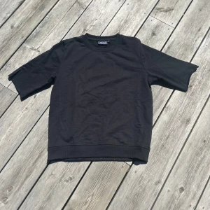 Svart t-shirt från OTP Leisurewear - En svart t-shirt från OTP Leisurewear i klassisk modell med rund hals och råa kanter på ärmarna. T-shirten är i bomull och har en loose fit som ger en avslappnad vibe. Perfekt för dig som gillar enkel och stilren look. 