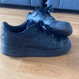 Svarta Nike Air Force 1 sneakers - Klassiska helsvarta Nike Air Force 1 sneakers i skinn med lågt skaft och svart sula. Skorna har rund tå, snörning och diskret Nike-logga på sidan och hälen. Perfekta för dig som gillar en clean och tidlös streetstil.