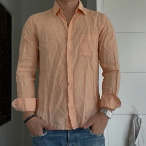 Persikofärgad linneskjorta Massimo Dutti - Snygg persikofärgad skjorta från Massimo Dutti i 100% linne. Skjortan har klassisk krage, bröstficka och knäppning framtill. Långärmad och perfekt för varmare dagar när du vill ha en chill och stilren look.