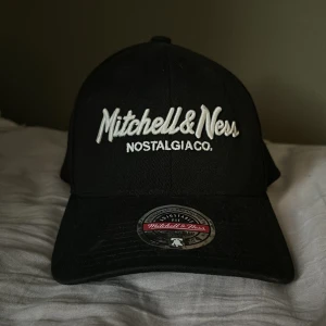 Svart keps från Mitchell & Ness - Svart keps från Mitchell & Ness med broderad vit logga framtill och baktill. Klassisk böjd skärm och justerbar snapback-spänne bak. Snygg streetstil och passar perfekt till en avslappnad look. Tillverkad i bomull.