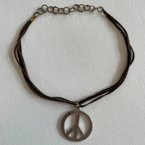 Halsband med peace-märke i silver - Coolt halsband med ett stort peace-märke i silverfärgad metall. Hängsmycket sitter på ett mörkbrunt band med flera trådar och justerbar kedja. Perfekt accessoar för dig som gillar retro eller boho-stil.
