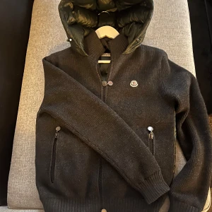 Tricot Cardigan Moncler (Svart stickad pufferjacka från Moncler) - Snygg svart pufferjacka från Moncler med stickade ärmar och framsida, samt glansig quiltad rygg och huva. Jackan har dragkedja framtill, två fickor med dragkedja och Moncler-logga på bröstet. Perfekt för kyliga dagar när du vill vara både varm och stilren. Digitalt kvitto finns!