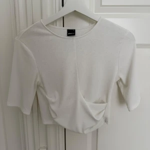 Vit omlottopp från Gina Tricot - Säljer en stilren vit topp från Gina Tricot med omlottdetalj framtill. Toppen har korta ärmar och en lätt croppad passform. Perfekt att matcha med jeans eller kjol för en clean look.