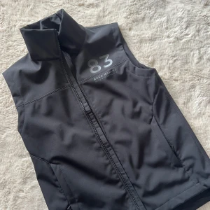 Svart väst från Race Marine XS - Sportig svart väst från Race Marine i storlek XS. Västen har hög krage, hel dragkedja framtill och två fickor med dragkedja. Utsidan är slät och insidan är fodrad med mjuk fleece för extra komfort. Snyggt tryck med siffran 83 på bröstet.