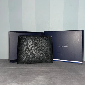 Tommy Hilfiger plånbok - Snygg svart plånbok från Tommy Hilfiger i äkta skinn med präglat geometriskt mönster på utsidan och diskret logga. Flera kortfack och stilren insida. Den ingår boxen från Tommy Hilfiger.