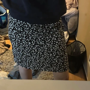 Svart blommig kortkjol - Supersöt svart kortkjol med vita små blommor över hela tyget. Kjolen har en rak passform och är perfekt för dig som gillar en enkel men ändå trendig look. Tyget känns lätt och flowy, perfekt för varmare dagar.