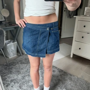 Blå jeanskjol från Diesel - Supersnygg blå jeanskjol från Diesel med cool omlott-look framtill och markerade sömmar. Kjolen har fickor både fram och bak samt bälteshällor. Perfekt för dig som gillar en trendig och avslappnad stil. Låg midja och kort modell.