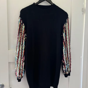 Svart klänning med paljettärmar Ciao Milano - Svart stickad klänning från Ciao Milano med långa ärmar täckta av färgglada paljetter i ränder. Klänningen har en hög, ribbad halsringning och en loose passform. 