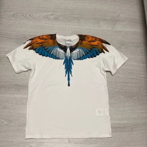 Marcelo Burlon  - Säljer en vit Marcelo Burlon t shirt med ikoniskt vingtryck i orange, blått och svart över axlar och bröst. T-shirten har rund halsringning och korta ärmar. Materialet är mjuk bomull och passformen är normal. Perfekt statement-plagg för dig som gillar unika prints.