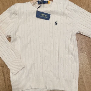 Vit kabelstickad tröja Polo Ralph Lauren - Vit kabelstickad tröja från Polo Ralph Lauren med klassisk rund halsringning och broderad marinblå logga på bröstet. Tröjan har lång ärm och ribbade muddar för en snygg passform. Perfekt för dig som gillar stilren och tidlös design.