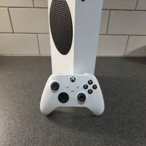 Xbox series s - Bilderna visar en Xbox Series S spelkonsol med tillhörande handkontroll och strömkabel. Konsolen har 512 GB SSD-lagring och är perfekt för gaming med vänner eller solo. Snygg, kompakt design som passar i alla rum. Originalförpackning ingår.