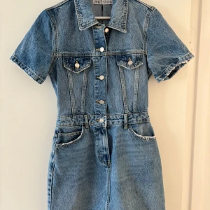 Jeansklänning från Zara i denim - Cool kort jeansklänning från Zara i klassisk blå denim. Klänningen har korta ärmar, krage, knappar framtill och fickor både fram och bak. Snygga sömdetaljer och bälteshällor i midjan ger en trendig look. Perfekt för dig som gillar streetstyle.