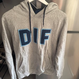 Grå DIF hoodie med blå bokstäver - Säljer en grå hoodie från Djurgårdens IF med stora blå DIF-bokstäver framtill. Tröjan har huva med mörkblå snören och en klassisk känguruficka. Mjuk och skön bomullsblandning.