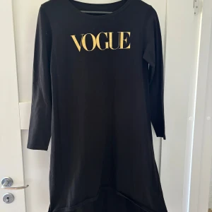 Svart klänning  - Svart långärmad klänning med guldig VOGUE-text framtill. Rak modell med rund halsringning och något längre baktill. Mjuk och bekväm