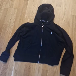 Svart hoodiejacka från Polo Ralph Lauren - Svart hoodiejacka från Polo Ralph Lauren med dragkedja framtill och klassisk logga broderad på bröstet. Jackan har huva men saknqr snöre till snörning och har två fickor framtill. Perfekt för en chill och stilren look. Använd fåtal gånger.