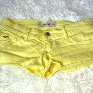 Gula jeansshorts från Hollister - Snygga gula jeansshorts från Hollister med råa kanter och klassisk femficksdesign. Shortsen har låg midja, knapp och dragkedja framtill samt Hollisters logga broderad på fickan. Perfekta för soliga dagar och en chill stil.