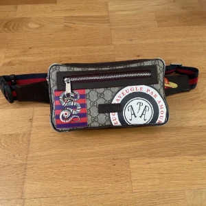  axelväska med tryck och dragkedja - Cool axelväska med patchar med ormmotiv och text, samt rödblå randig axelrem. Väskan har en dragkedja framtill och detaljer i brunt skinn. Perfekt för dig som vill sticka ut med en unik accessoar.