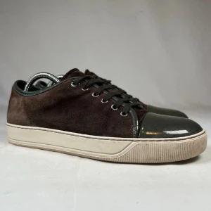 Bruna Lanvins - Snygga bruna sneakers i mocka med cool lackad mörkgrå tå och matchande snörning. Skorna har en vit platt sula och detaljer i mörkgrått runt öppningen. Perfekta för dig som gillar en stilren men unik look. Skorna är det ända som ingår