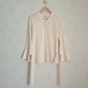 Carin Wester krämvit blus - 🧁FÖRST TILL KVARN!🧁 En krämvit beige blus från märket Carin Wester. Fina ärmarna med knytband runt som man kan knyta till rosetter runt handleden om man vill. V-krage med fina resin knappar.