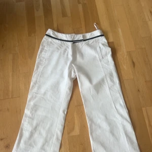 Vita linnebyxor med svart detalj - Snygga vita linnebyxor med rak passform och svart dekorativt band i midjan. Byxorna har fickor fram och en bakficka med knapp. Perfekta för en fräsch och avslappnad look under varmare dagar.