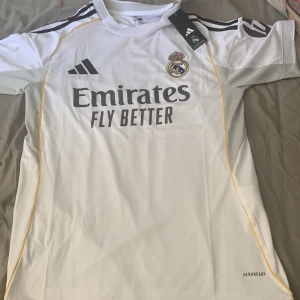 Real Madrid Adidas fotbollströja vit - Säljer en officiell Real Madrid fotbollströja från Adidas i vitt med svarta detaljer och gula linjer. Tröjan har klubbmärke på bröstet, Adidas-logga och sponsortryck. Tillverkad i lätt och ventilerande material (AEROREADY). Perfekt för dig som älskar fotboll och vill bära klubbens färger.