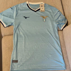 Ljusblå Lazio fotbollströja Mizuno - Ljusblå officiell SS Lazio fotbollströja från Mizuno med klubbmärke på bröstet och mörkblå detaljer vid krage och ärmslut. Tröjan har korta ärmar och är tillverkad i ett lätt, ventilerande material perfekt för match eller träning.