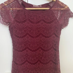 Vinröd spetstopp med kort ärm - Vinröd topp i spets med vackert mönster och korta ärmar. Toppen har en rund halsringning och är figurnära. Perfekt för dig som vill ha en romantisk och trendig look. Materialet är mjukt och följsamt.