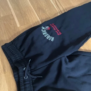 Svarta mjukisbyxor från Hoodrich barnstorlek S 8-10 års ålder  - Svarta mjukisbyxor från Hoodrich med broderad logga och texten 'From Nothing To Something' i vitt och rött på ena benet. Byxorna har resår och snörning i midjan för skön passform. Perfekta för chill eller streetwear.