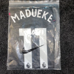 Chelsea Madueke #11 Nike fotbollströja - Chelsea Madueke #11 Premier League tröja från Nike. Svart bas med grått mönster, rosa och gula detaljer på ärmsluten. Broderad Chelsea-lejonlogga och Nike Swoosh på bröstet. Tillverkad i lätt och ventilerande material, perfekt för match eller träning. Storlek S