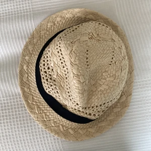 Stråhatt från Kappahl med svart band - Snygg stråhatt från Kappahl i naturfärgat flätat material med ett svart band runt kullen. Perfekt för soliga dagar och ger en chill vibe till din outfit. Hatten har klassisk form och är luftig tack vare det öppna flätmönstret.