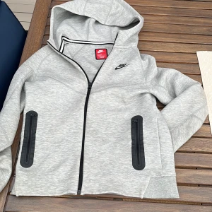 Grå hoodiejacka från Nike - Snygg grå hoodiejacka från Nike med svart logga på bröstet. Jackan har hel dragkedja, två svarta fickor med dragkedja och huva. Perfekt för chill dagar eller när du vill ha en sportig look. Mjuk och skön i bomullsmix.