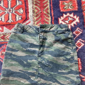 Camouflagekjol från Zadig & Voltaire - Cool kortkjol från Zadig & Voltaire i camouflage-mönster med gröna och beige toner. Kjolen har bälteshällor, knapp och dragkedja framtill samt bakficka med knapp och broderad Zadig Sisters-detalj.Aldrig använd  