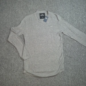 Grå stickad tröja från Hollister - Snygg grå stickad tröja från Hollister med rund halsringning och diskret logga broderad på bröstet. Långärmad modell i mjukt material som passar perfekt till jeans eller chinos. Enkel och stilren design som funkar året runt.