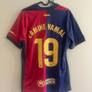FC Barcelona Lamine Yamal matchtröja Nike - Snygg FC Barcelona matchtröja med Lamine Yamal och nummer 19 på ryggen. Tröjan är röd och blå, har korta ärmar och är tillverkad i ett lätt, ventilerande material. Nike-logga, klubbmärke och sponsorer i guld på bröstet. Perfekt för dig som älskar Barca!