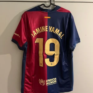 FC Barcelona Lamine Yamal Cactus Jack tröja - Säljer en FC Barcelona matchtröja från Nike med Lamine Yamal och nummer 19 på ryggen. Tröjan har klassisk halva-röd halva-blå design, guldiga detaljer och Cactus Jack-grafik framtill. Broderat klubbmärke och sponsorer på ärm och rygg. Tillverkad i lätt och ventilerande polyester.
