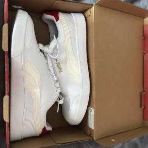 Vita sneakers från Puma med röda detaljer - Helt nya inte ens andvända box medföljer vid köp. Fräscha vita sneakers från Puma med klassisk design, guldfärgad logga på sidan och röda detaljer vid hälen. Ovandel i syntet, foder i textil och sula i gummi. Perfekta för dig som gillar stilrena och sportiga skor med modern vibe.