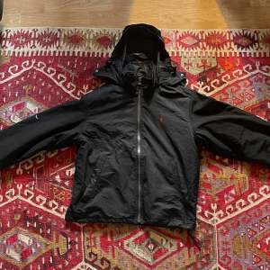 Svart vindjacka från Polo Ralph Lauren - Svart vindjacka från Polo Ralph Lauren med röd logga på bröstet. Man kan vika ut och in huvan beroende på väder, dragkedja hela vägen och två fickor framtill. Tillverkad i 100% polyester och har en klassisk, lite loose passform. Perfekt för blåsiga dagar.