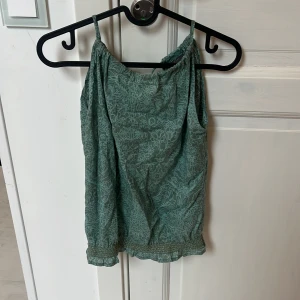 Grön blommig linnetopp Filippa K - Supersöt grön linnetopp från Filippa K med tunna axelband och blommigt mönster. Toppen har v-ringning i ryggen och smockad kant nertill för en snygg passform. Perfekt för varma dagar och enkel att styla med jeans eller kjol.