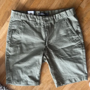 Olivgröna shorts från Dressmann oanvända - Snygga olivgröna shorts från Dressmann med klassisk passform. De har två sidfickor, två bakfickor med knapp och hällor för bälte. Tillverkade i mjuk bomull som känns skön mot huden. Perfekta för varma dagar när du vill ha en clean och stilren look.