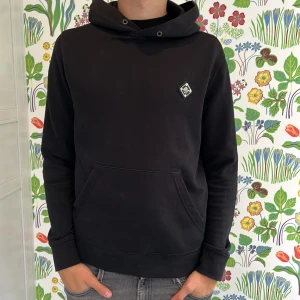 Svart hoodie  - Svart hoodie från J- Lindeberg med klassisk känguruficka och en liten logopatch på bröstet. Tröjan är tillverkad i mjuk bomullsmix. Storleken är S och sitter som S. sicket är bra inga defekter förutom att snörena är borta.