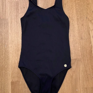Svart ribbad baddräkt från KappAhl - Snygg svart baddräkt från KappAhl i storlek 134/140. Modellen är enkel och stilren med ribbad struktur och ett litet guldfärgat emblem framtill. Perfekt för strand eller poolhäng hela sommaren.