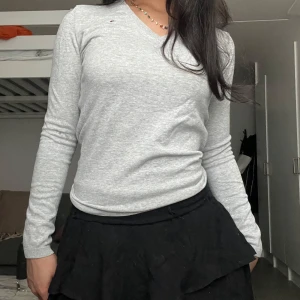 Grå v-ringad tröja Tommy Hilfiger - Snygg ljusgrå v-ringad tröja från Tommy Hilfiger med liten broderad logga på bröstet. Tröjan är långärmad och har en klassisk, normal passform. Perfekt att styla med kjol eller jeans för en clean look.