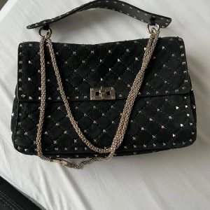 Valentino Garavani rockstund spike bag - Säljer en svart Valentino väska med coola guldfärgade nitar och kedjerem. Väskan har en snygg låsdetalj framtill och är gjord i mjuk mocka. Perfekt för dig som gillar edgy vibes och vill sticka ut med accessoarerna. Original och kvitto finns. 