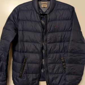 Blå pufferjacka från Hilfiger Denim - Säljer en mörkblå pufferjacka från Hilfiger Denim med quiltad design och dragkedja framtill. Jackan har svarta detaljer vid fickorna och en liten broderad logga på bröstet. Perfekt för kyliga dagar. Mindre skavanker på kanten på fickorna förekommer (se bild)